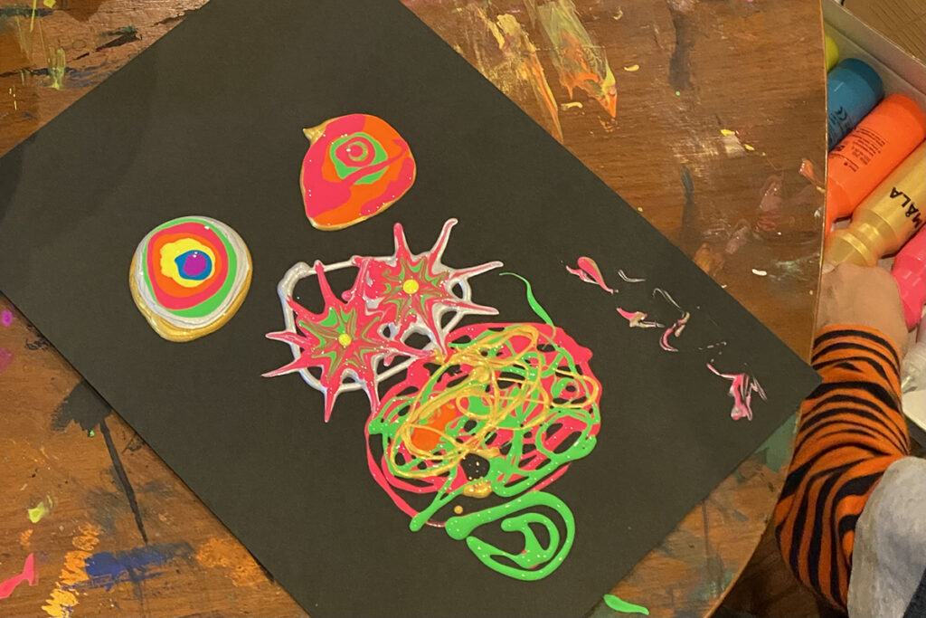 黒い画用紙の上に、MÅLA絵の具で描かれた鮮やかな模様。右端には、絵の具を手に取る子どもの手が写っている。