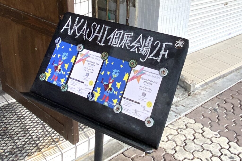 AKASHI初個展の会場案内板。黒板に“AKASHI個展会場2F”とチョークで書かれ、フライヤーが貼られている