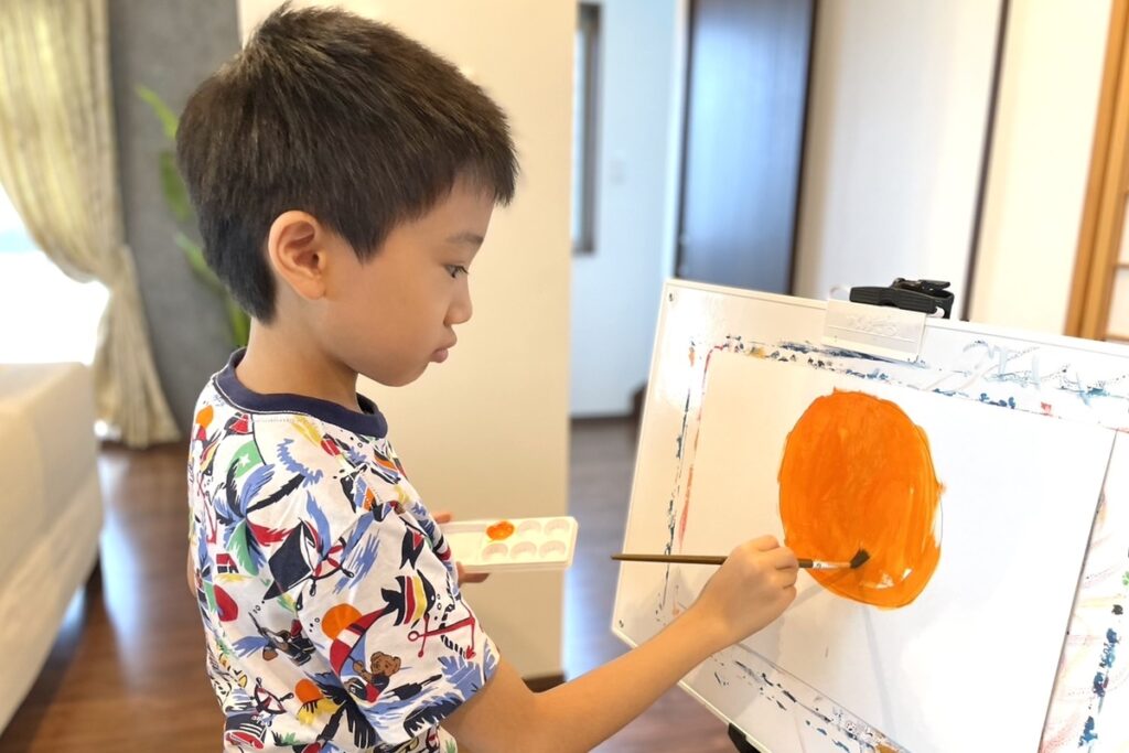 真剣な表情でキャンバスに向かい、オレンジの夕日の絵を描くAKASHI