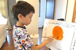 真剣な表情でキャンバスに向かい、オレンジの夕日の絵を描くAKASHI