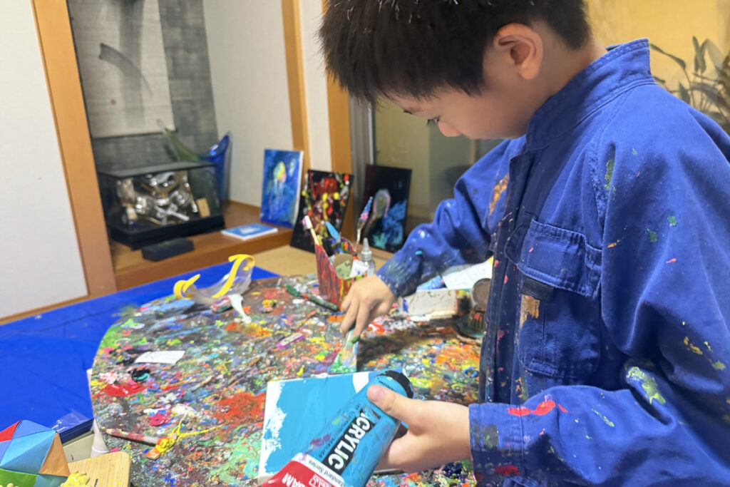 青い作業着を着て、キャンバスに絵の具を塗るAKASHI。制作に夢中になる姿。