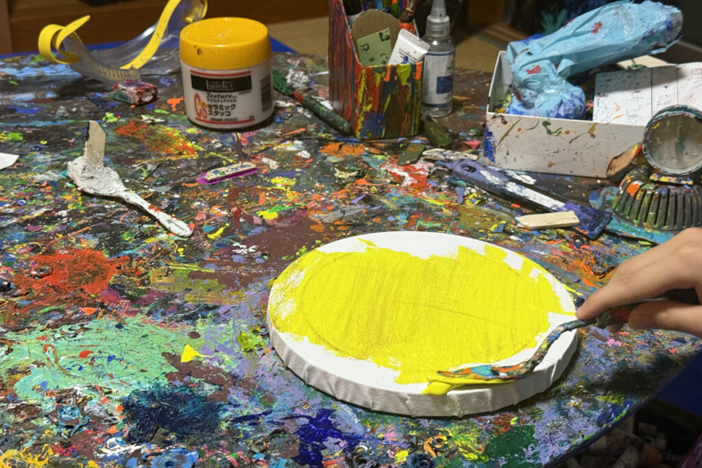 絵を描き始めるAKASHIの制作風景。黄色い絵の具をパレットナイフで丸いキャンバスに塗り重ねている様子。