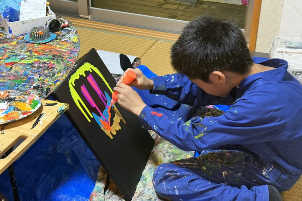 黒いキャンバスに蛍光オレンジ、ピンク、ブルーを重ねながら描くAKASHI(9歳)の制作風景。色が垂れて混ざり合い始めている。