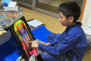 蛍光ピンクやオレンジ、ブルーの絵の具が垂れ、黒いキャンバスにカラフルな模様が広がる。
AKASHI(9歳)が笑顔で指を差しながら描いている様子。