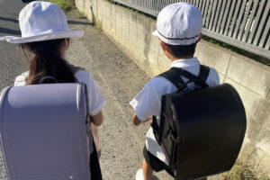 新しい制服とランドセルで登校するAKASHIとRIRIの後ろ姿。転校初日の朝の風景。