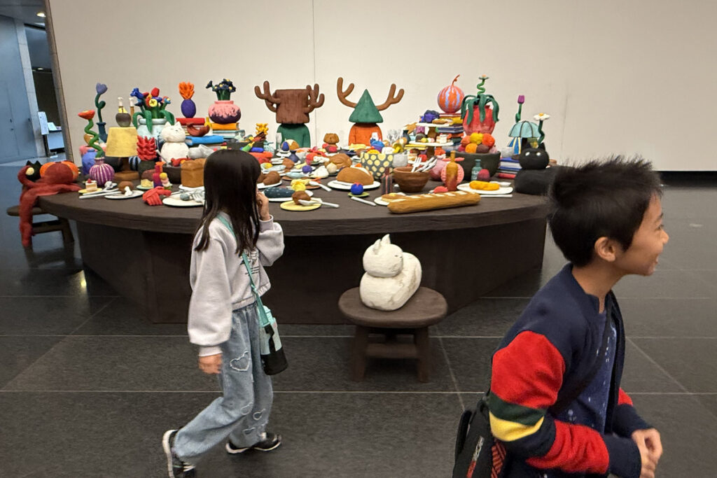 平子雄一展の会場で、カラフルな立体作品を前に歩くAKASHIとRIRI。色とりどりの食べ物のような作品が並ぶ。