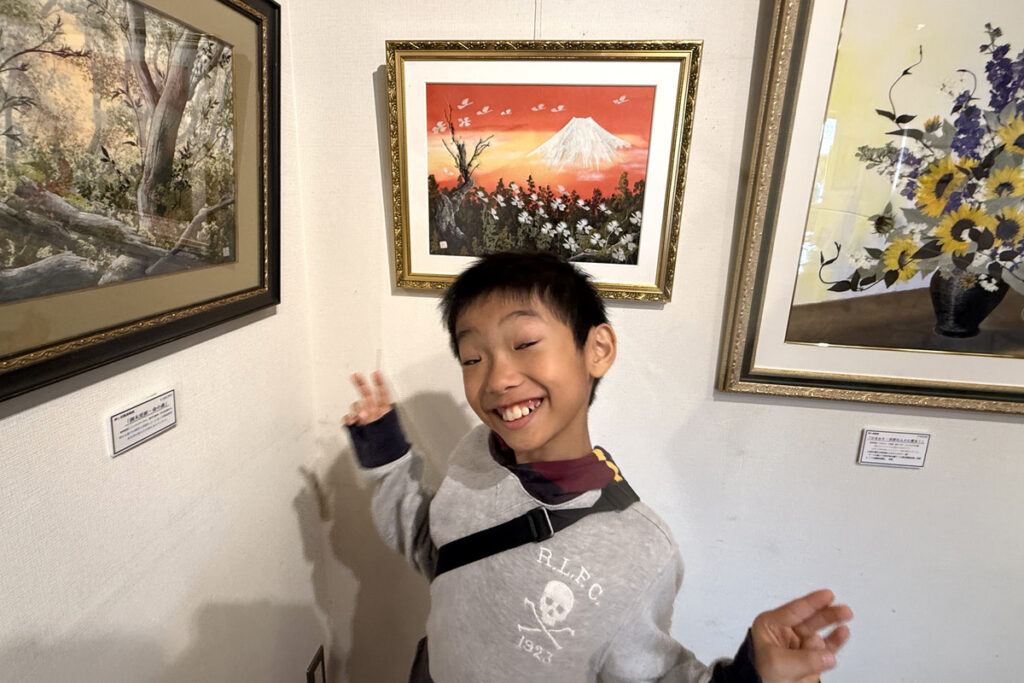 山本和子さんの富士山の押し花作品の前で笑顔を見せるAKASHI。倉敷・ギャラリー十露にて。