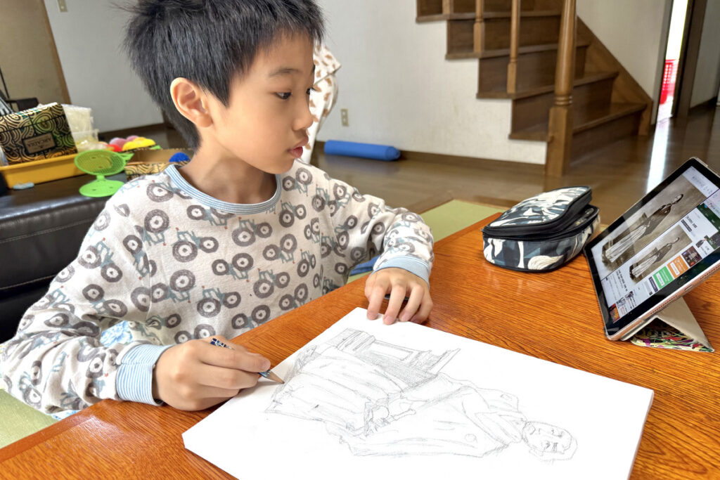 坂本龍馬の写真を見ながら下描きをするAKASHI。タブレットを横に置き、集中して鉛筆を走らせている。