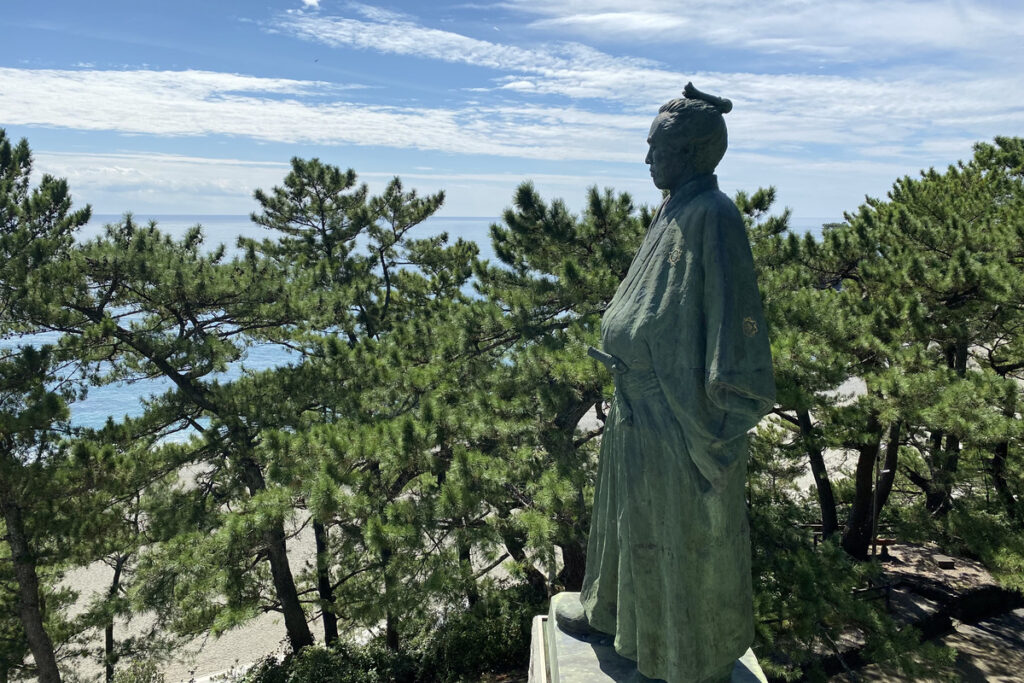 高知・桂浜に立つ坂本龍馬像。青空と松林を背に、遠くを見つめている姿。